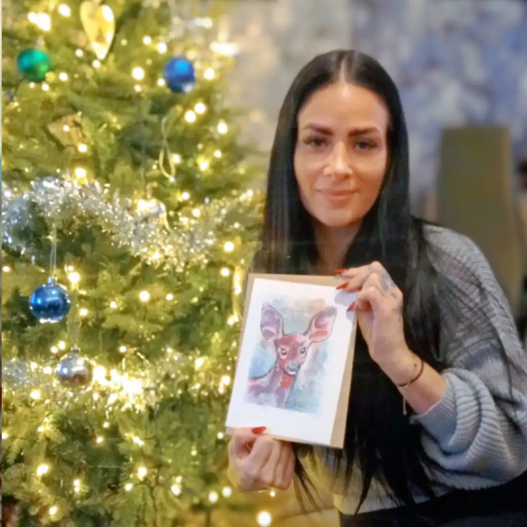 An image of Lauren W, a customer, enjoying her free print of a watercolour female deer portrait . She is smiling in her living room next to her Christmas tree, holding up the small hand signed and numbered watercolour print and the certificate of authenticity from Jamie C. Goodwin available at JamieHasWings.com
