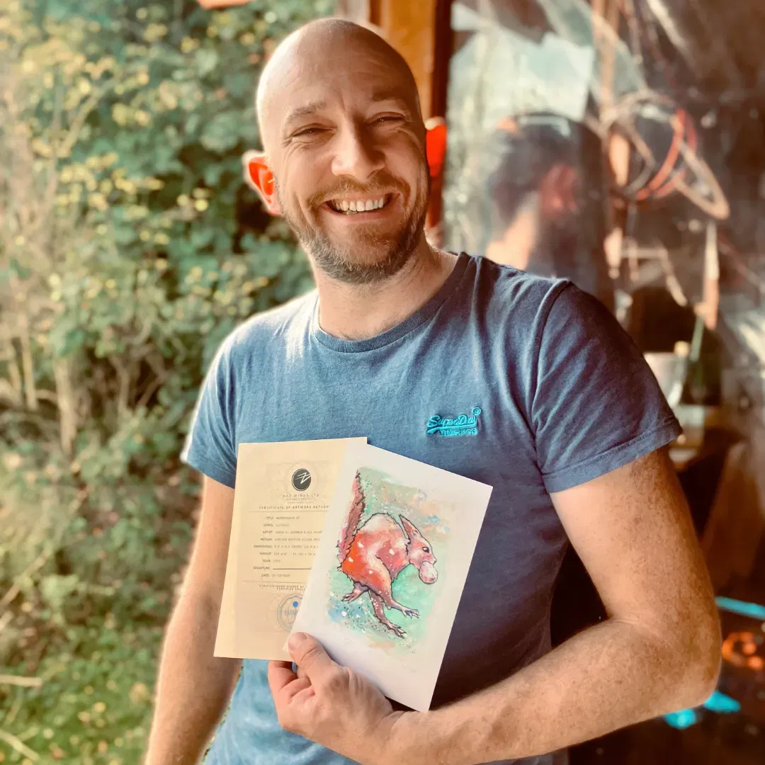 An image of Adam B, a customer, enjoying his free print of a watercolour red squirrel . He is smiling in the doorway of Jamie's Cabin, holding up the small hand signed and numbered watercolour print and the certificate of authenticity from Jamie C. Goodwin available at JamieHasWings.com