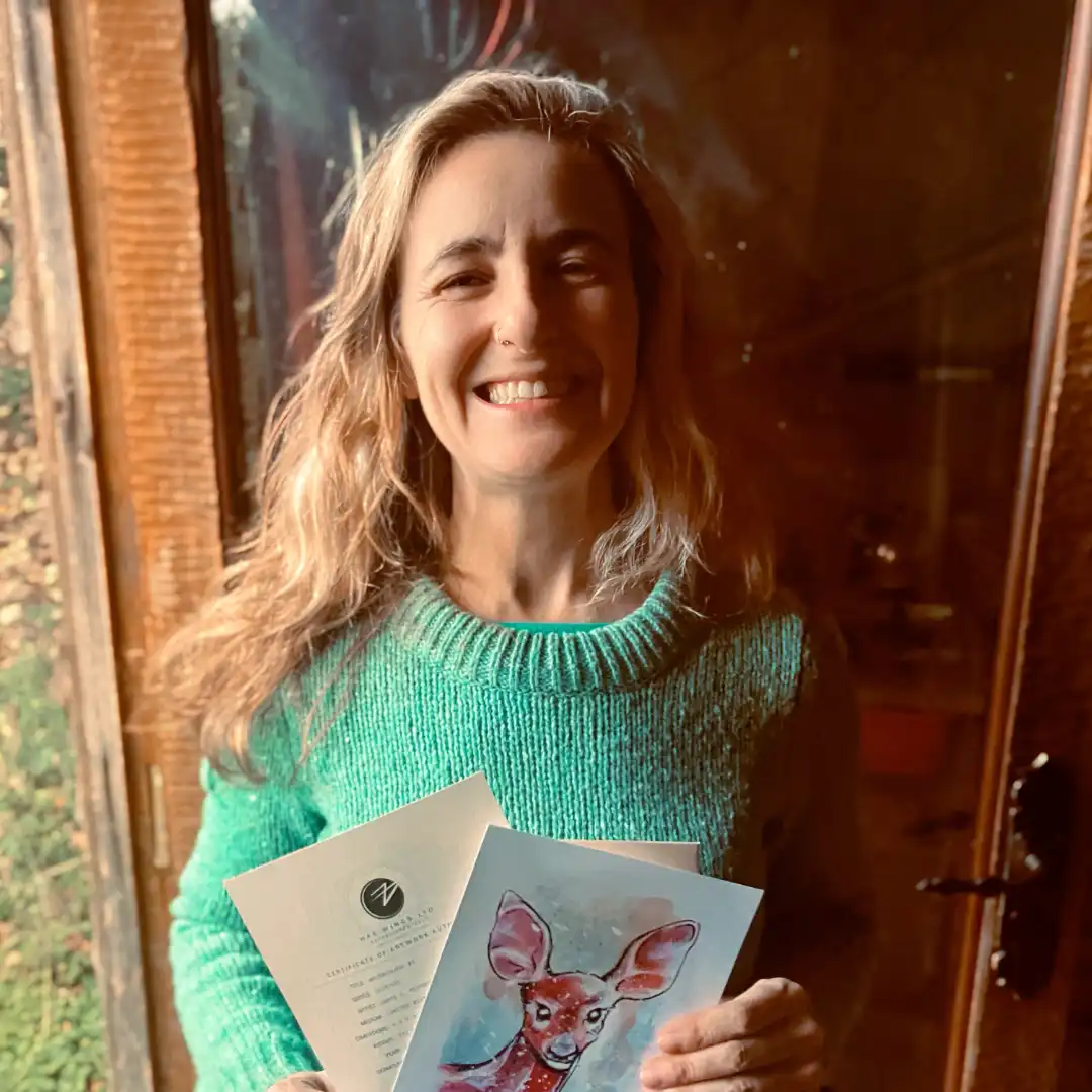 An image of Belén P.H., a customer, enjoying her free print of a watercolour female deer portrait . She is smiling in the doorway of Jamie's Cabin, holding up the small hand signed and numbered watercolour print and the certificate of authenticity from Jamie C. Goodwin available at JamieHasWings.com