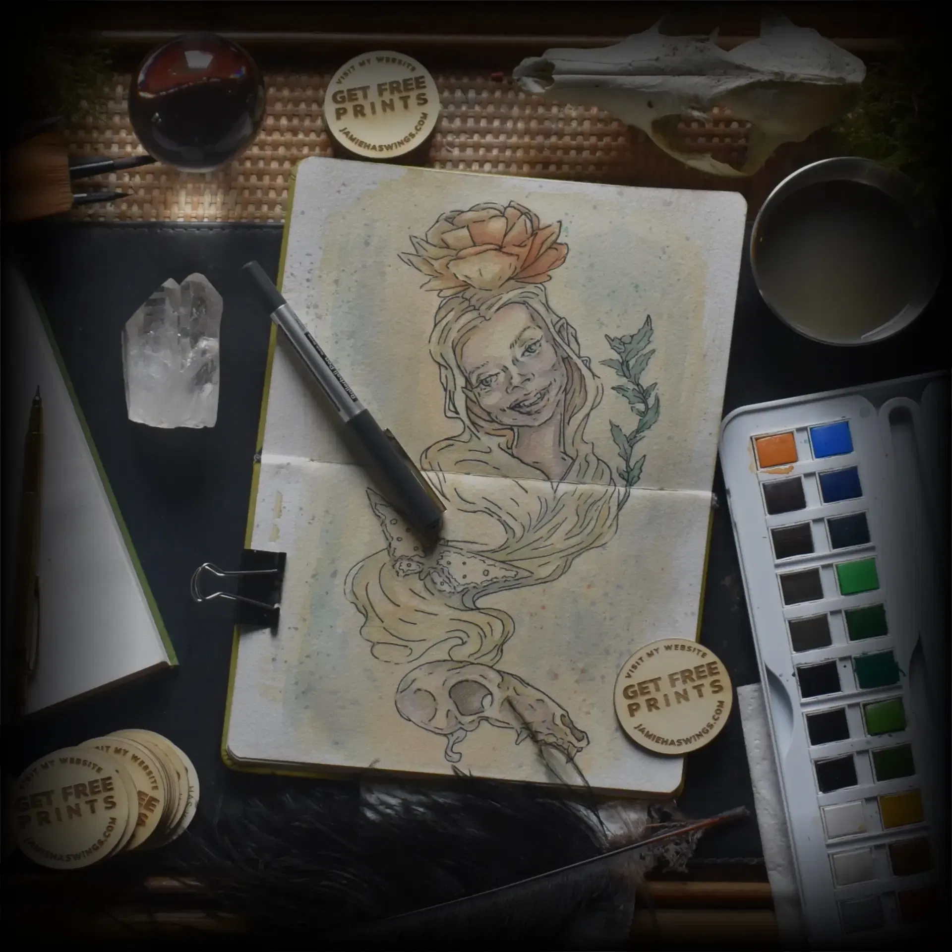 A snapshot of Jamie C. Goodwin's art studio to help communicate Jamie C. Goodwin's Artist Statement. The photograph is showing the top of his bamboo desk covered in watercolour paints, animal skulls, crystals, rocks, and sketchbooks showing his art. The picture also shows his wood round business card which says visit my website get free prints jamiehaswings.com