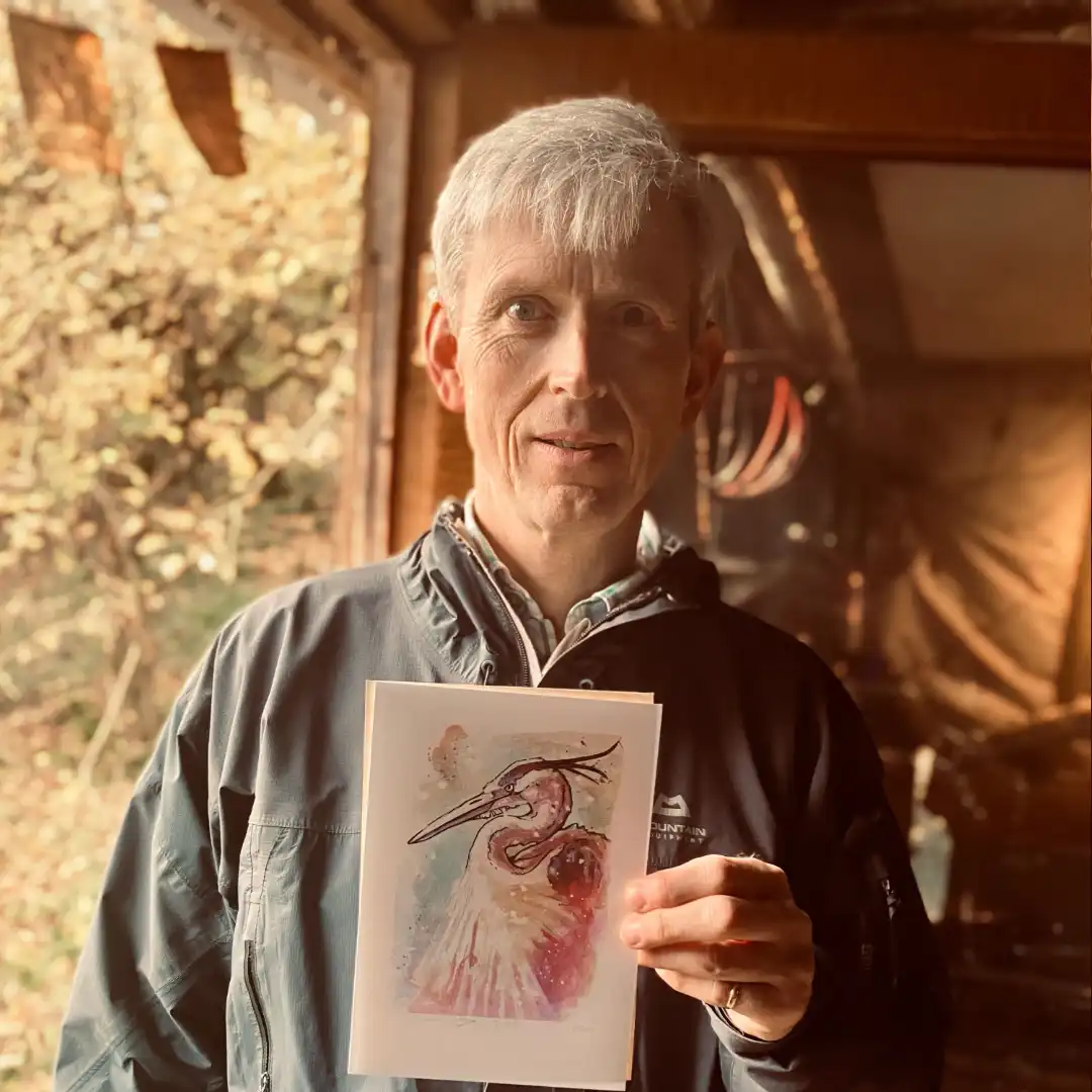An image of James H., a customer, enjoying his free print of a watercolour heron. She is smiling in the doorway of Jamie's Cabin, holding up the small hand signed and numbered watercolour print and the certificate of authenticity from Jamie C. Goodwin available at JamieHasWings.com
