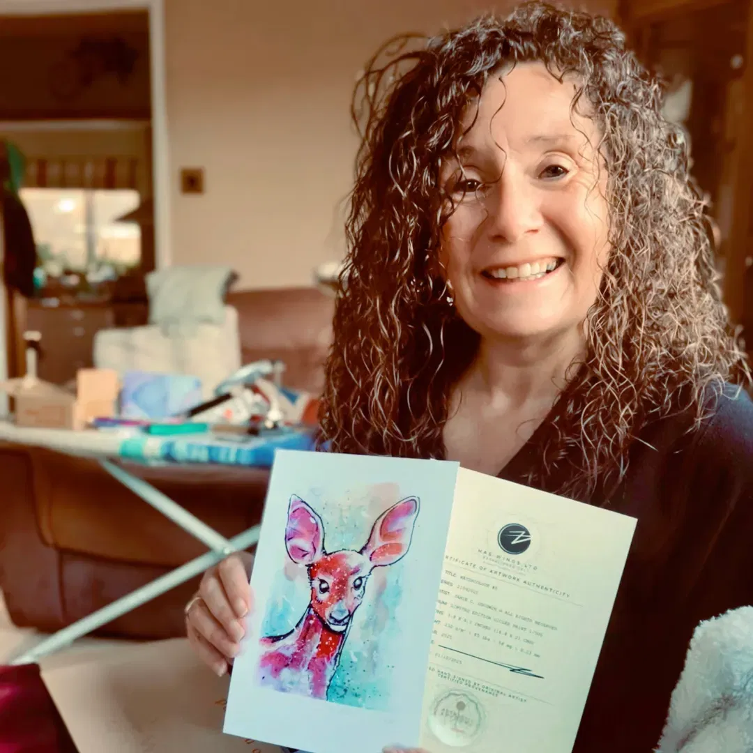 An image of Sam W, a customer, enjoying her free print of a watercolour female deer portrait . She is smiling in her living room, holding up the small hand signed and numbered watercolour print and the certificate of authenticity from Jamie C. Goodwin available at JamieHasWings.com