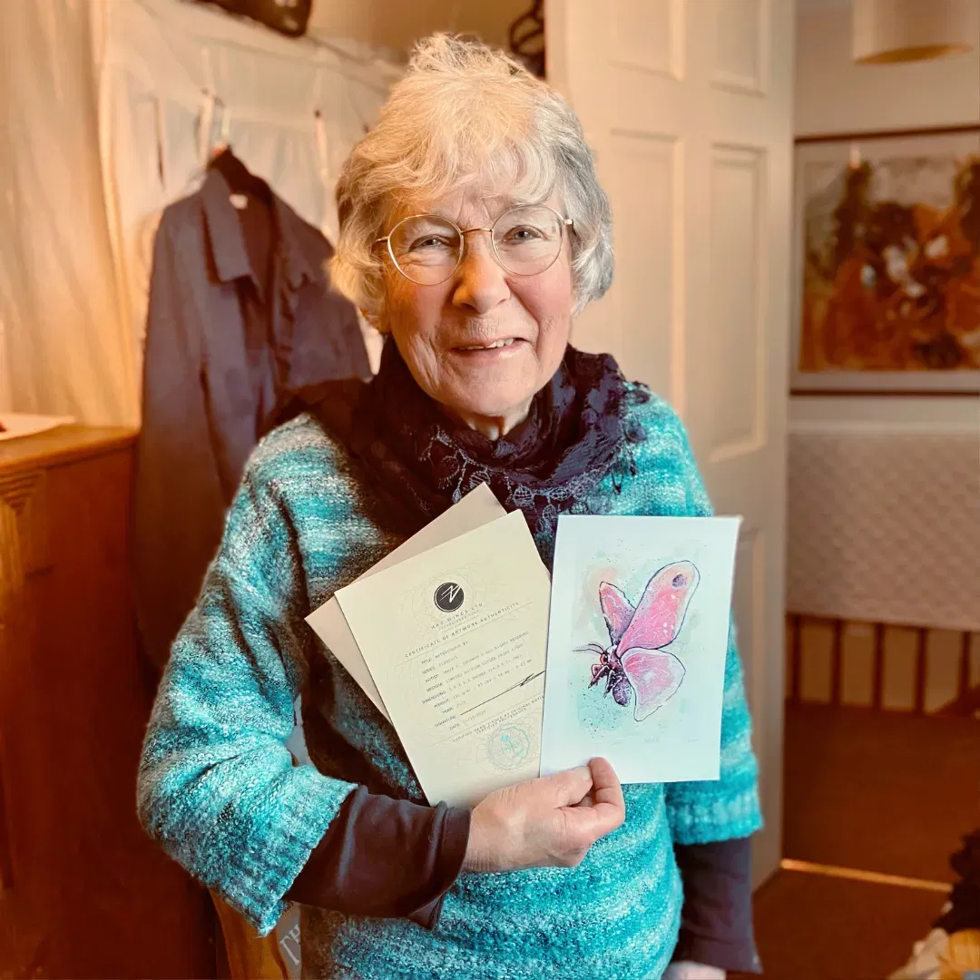 An image of Jane B, a customer, enjoying her free print of a watercolour moth. She is smiling in her home, holding up the small hand signed and numbered watercolour print and the certificate of authenticity from Jamie C. Goodwin available at JamieHasWings.com