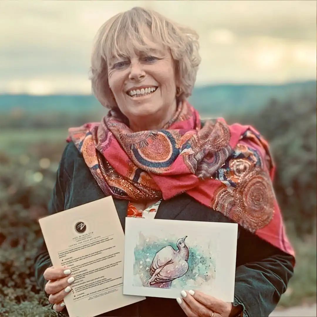 An image of Caroline H, a customer, enjoying her free print of a watercolour pigeon. She is smiling while standing outdoors in a beautiful valley, holding up the small hand signed and numbered watercolour print and the certificate of authenticity from Jamie C. Goodwin available at JamieHasWings.com