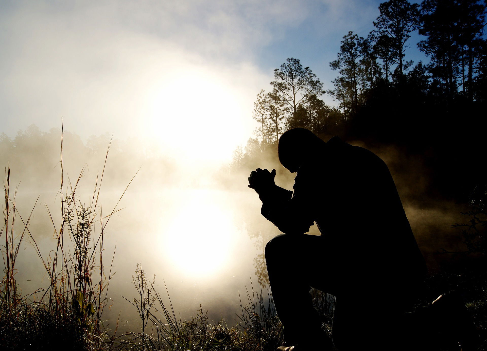 A silhouette of a person in prayer, kneeling with hands clasped against a backdrop of a misty, sunlit morning. The rising sun filters through the haze, enveloping the figure in a soft glow, creating an atmosphere of tranquility and devotion.